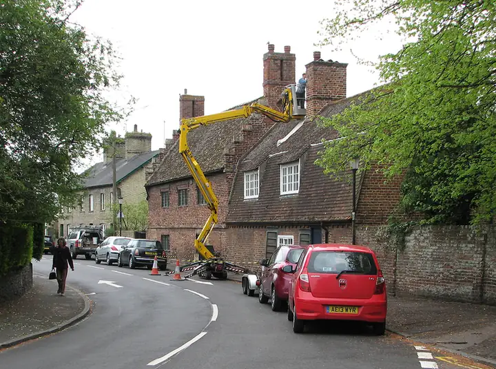 Roofer in a cherry picker repointing a tall brick chimney stack on an old tiled terrace beside a village road
