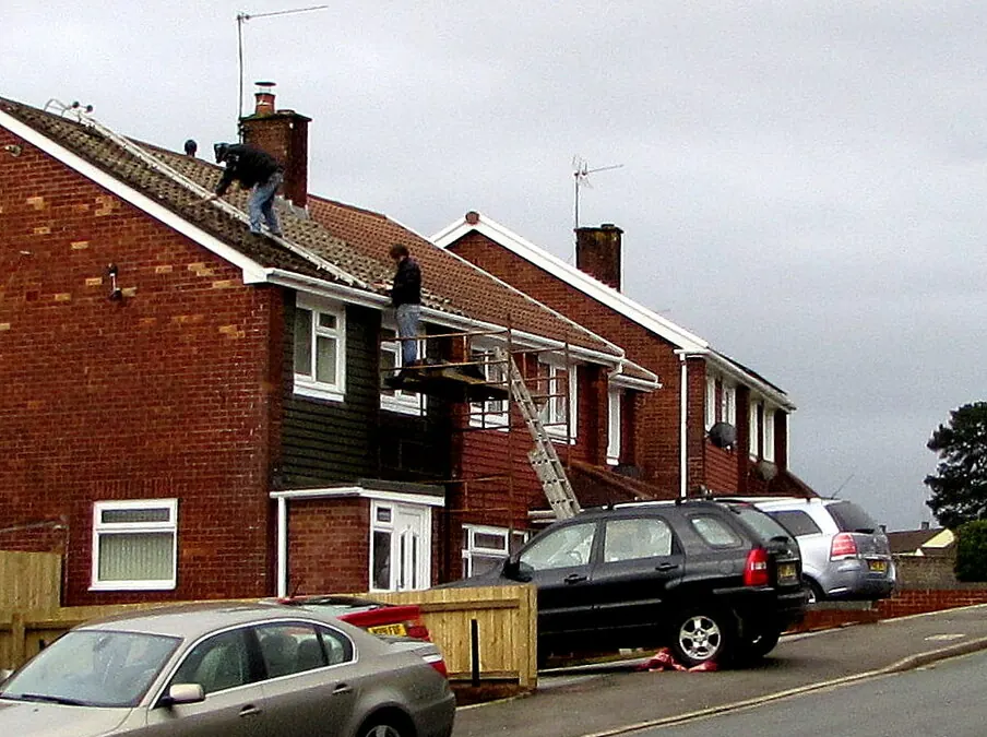 Two roofers working on the tiled roof of a red brick semi-detached house, one at the chimney and one on a ladder platform at the eaves