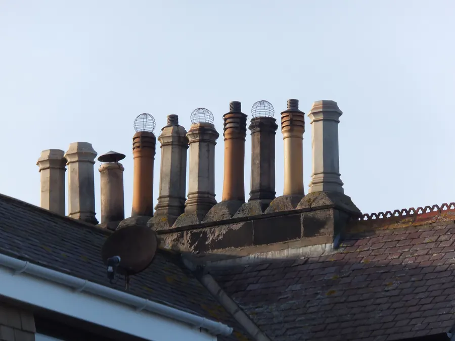 Long row of clay chimney pots, some fitted with wire bird guards and cowls, on a stone stack above a slate roof