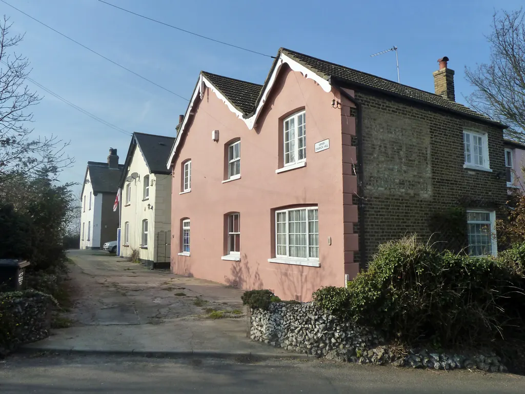 Pink-rendered and brick cottages with pitched roofs and chimneys behind a flint wall on Upper Church Hill, Stone